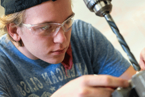 A white male student with blonde hair and wearing a black cap, glasses, and a powder-blue American Classic t-shirt, works on something that is out of focus at bottom left of frame. A drill is visible with a lengthy drill bit that extends down toward whatever he is holding in his fingers. The student’s bright blue eyes are visible as he focuses on his work.  