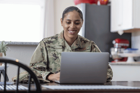 A Black woman wearing military fatigues sits and smiles in front of a laptop at a kitchen table in a residence.